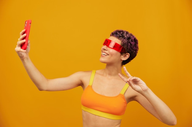 Woman blogger smiling in unusual millennial glasses taking selfies on her phone in sportswear against an orange studio backdrop free space