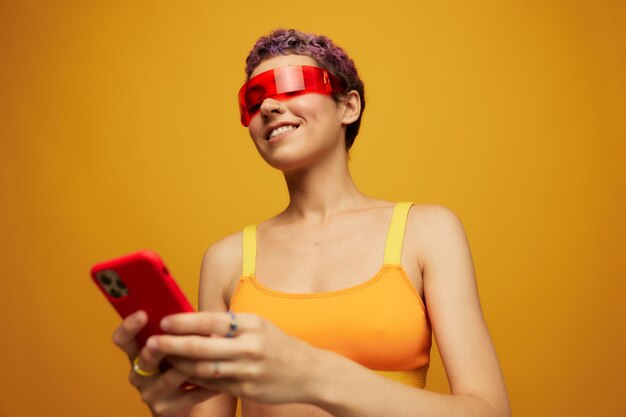 Woman blogger smiling in unusual red millennial glasses holding her phone and looking at the screen against an orange background in a stylish yellow top