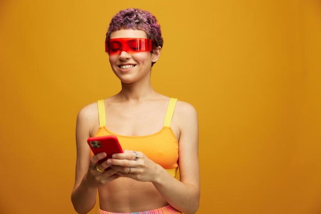 Woman blogger smiling in unusual red millennial glasses holding her phone and looking at the screen against an orange background in a stylish yellow top