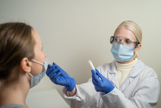 보호 안경을 쓴 금발의 여성 의사가 환자에게 코로나바이러스 검사를 합니다. 코로나19 팬데믹 기간 동안 건강을 보호합니다.