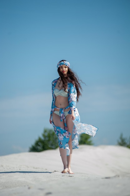 A woman in a blue bikini and a flowered skirt walks in the sand.