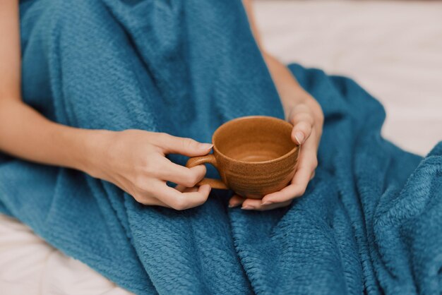 Woman in blue blanket with cup of coffee sitting on bed covered with blanket