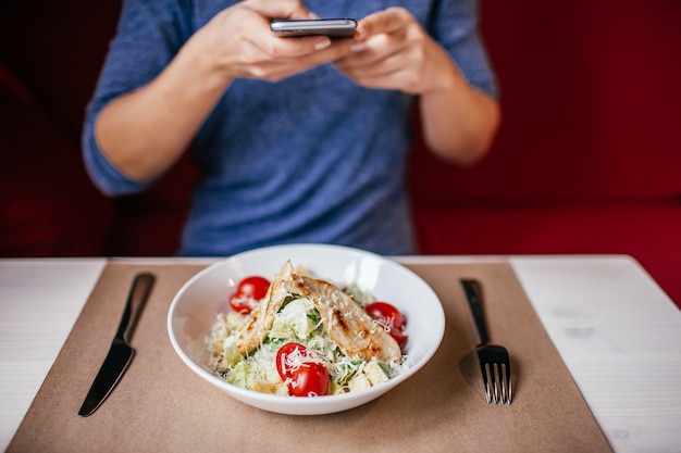 A woman in a blue blouse taking photo of fresh salad caesar on\
the table with her smartphone