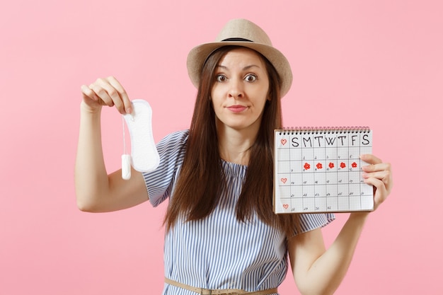 Woman in blue dress, hat holding sanitary napkin, tampon female periods calendar, checking menstruation days isolated on pink background. Medical, healthcare, gynecological choice concept. Copy space.