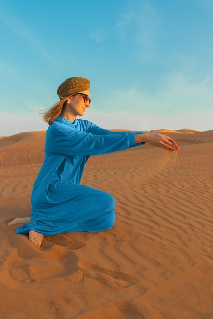 A woman in a blue dress sits in the desert with the sun setting behind her.