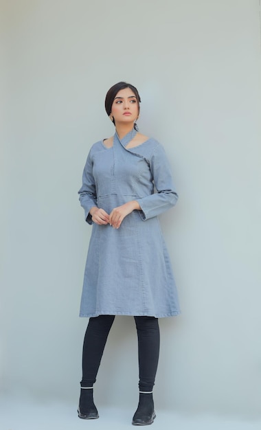 A woman in a blue dress stands against a white wall.