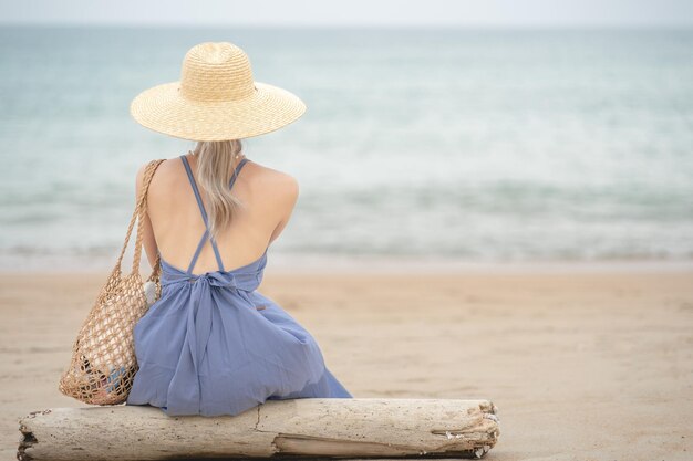 Woman in  blue dress and straw hat, sitting on a timber by the ocean.