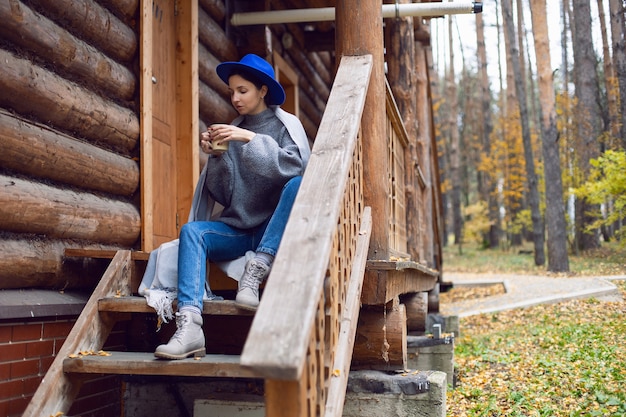 Женщина в синей шляпе, шарфе и кружке стоит у лестницы деревянного дома, в лесу осенним утром и думает о жизни