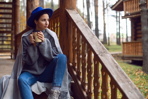 Woman in a blue hat and scarf and a mug stands at a wooden house in the woods