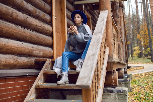 Woman in a blue hat and scarf and a mug stands at a wooden house in the woods