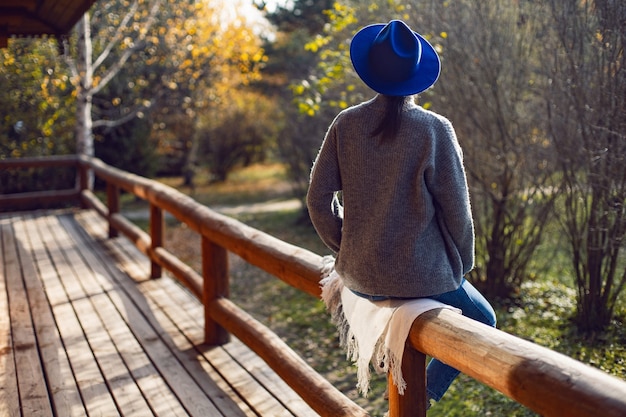 Woman in a blue hat and sweater is sitting back on wooden fence near wooden house made of logs in autumn