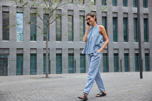  woman in blue outfit enjoys cellphone conversation in roaming while traveling abroad makes smartphone calling via app strolls outdoor near modern establishment