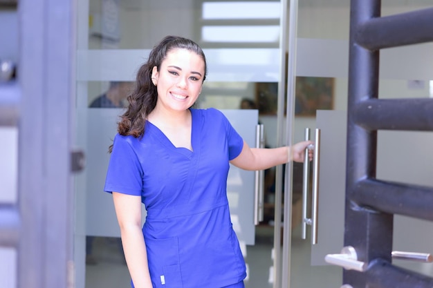 A woman in blue scrubs stands in front of a glass door.