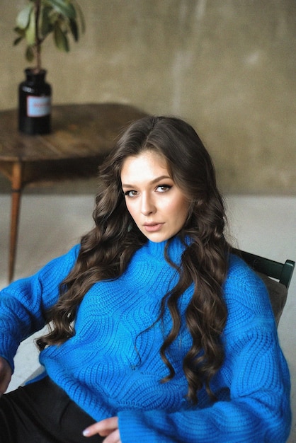 A woman in a blue sweater sits in front of a table with a bottle of wine on it.