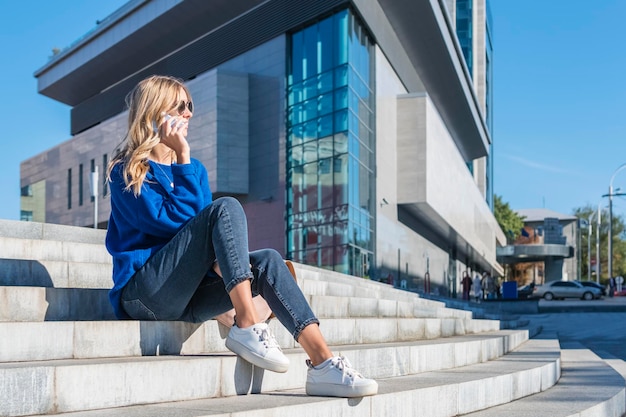 Donna in maglione blu che parla al telefono seduta sui gradini della città vicino all'edificio degli uffici