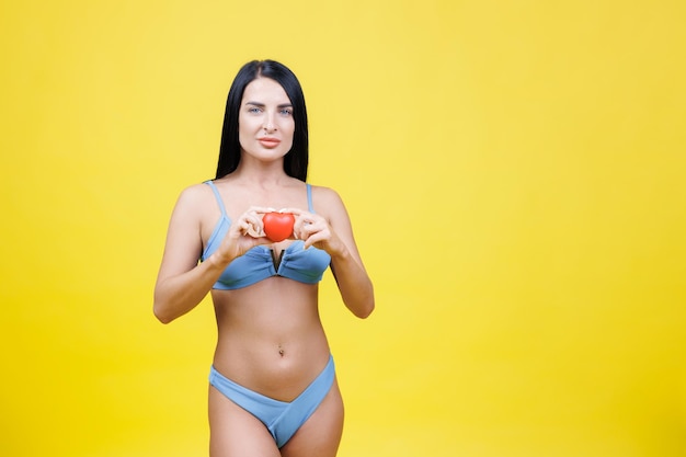 Woman in blue swimsuit holding little red heart in hand standing isolation in studio on yellow background Summer healthy rest concept