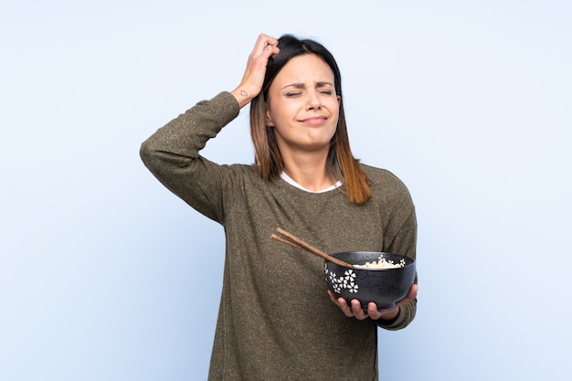 Женщина над синей стеной с сомнением и смущенным выражением лица, держа миску лапши с палочками для еды