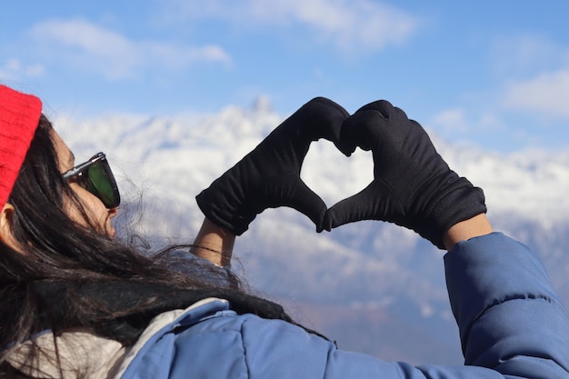 Photo a woman in a blue winter coat and gloves making a heart shape with her hands in front of a snowy mountain.