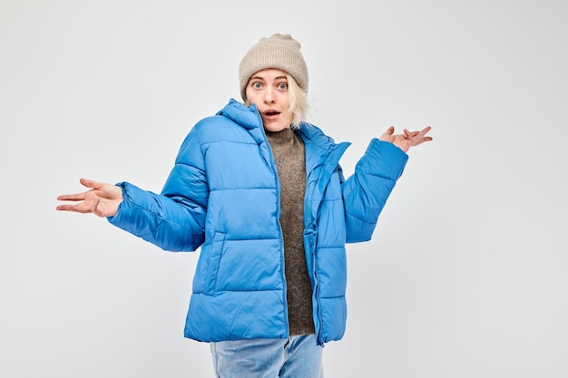 Woman in blue winter jacket and beanie shrugging with a puzzled expression on a white background