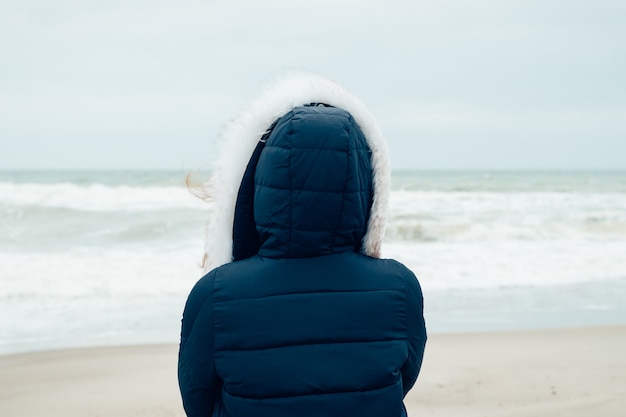 Woman in a blue winter jacket with a hood stands on the winter beach and looking at sea