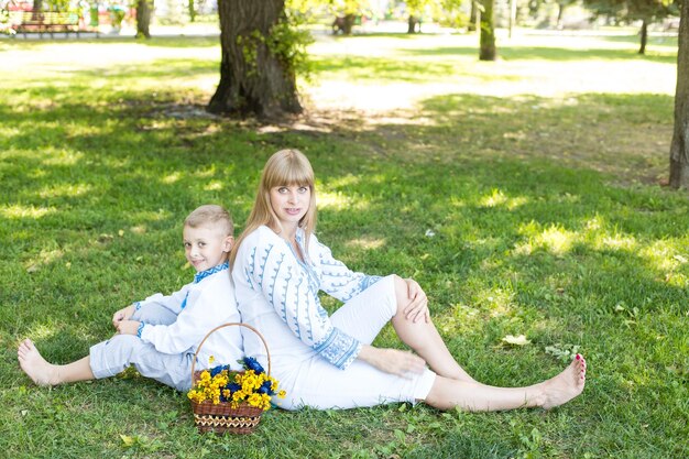 A woman and a boy sit on the grass with a basket full of flowers.