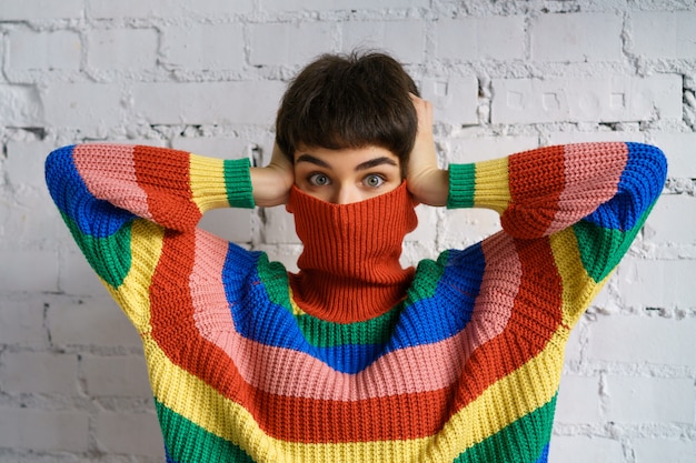 woman in a bright multicolored rainbow sweater hides her face and covers her ears with her hands.