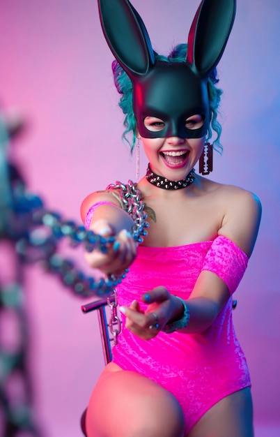 The woman in a bright pink bodysuit and rabbit mask poses against a bright background with a chain