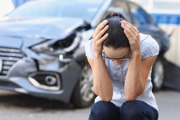 A woman at a broken car sits holding her head closeup blurry shock at the driver after an