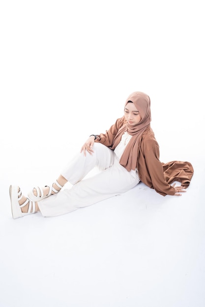 A woman in a brown hijab sits on the floor in a white background.