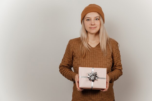 Woman in a brown sweater and hat holding a pink gift in her hands on a gray background