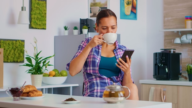 Woman browsing on smartphone while drinking green tea, in the morning during breakfast. Holding phone device with touchscreen using internet technology scrolling, searching on intelligent gadget.