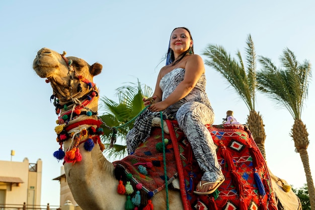 Woman in a brunette overalls riding a camel. Horizontal photo