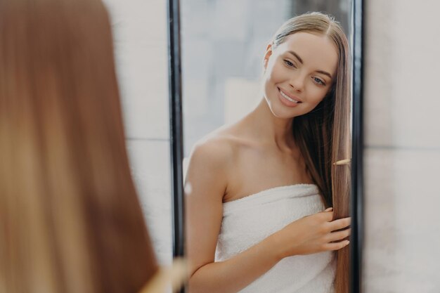 女性はバスルームでタオルに巻いた長い髪をブラシします。 ヘアケアと美容のコンセプト