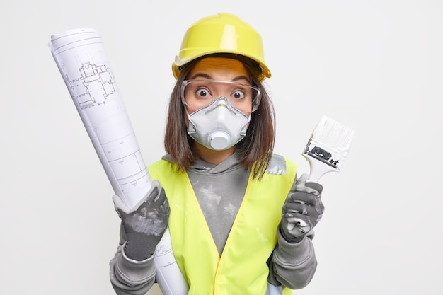 woman builder or engineer works on layout of room holds brueprint and paint brush wears working uniform and safety equipment busy with construction