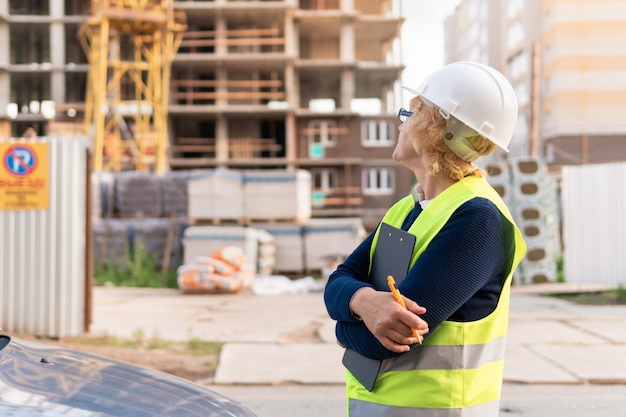 Woman Builder in a white helmet and yellow vest. Middle-aged woman working on a construction site. She's taking readings on a clipboard.