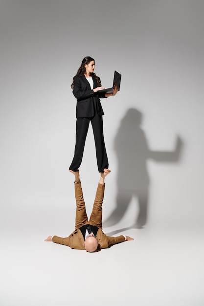 Woman in business attire with laptop balancing on feet of man formal wear couple of acrobats