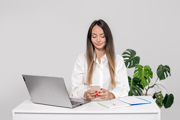 woman in business clothes sits at a table with a laptop and looks at the phone