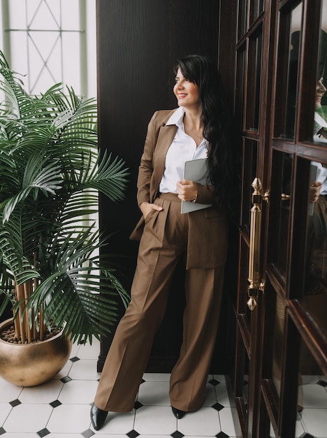 A woman in a business suit stands near the door in the office smiling holding a tablet