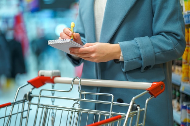 Foto compratore della donna con il carrello nella navata laterale del negozio con la lista della drogheria durante l'alimento di acquisto