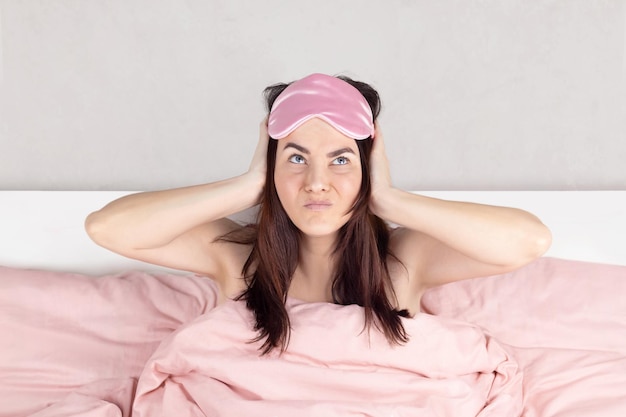 Woman cannot sleep she covers her ears with her hands the neighbors are noisy making repairs Pink sleep mask pink bed linen