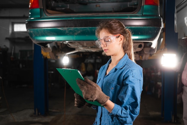 A woman car mechanic is wearing safety glasses and working overalls standing at a car service station holding a tablet in her hands and making notes about the necessary rebuild