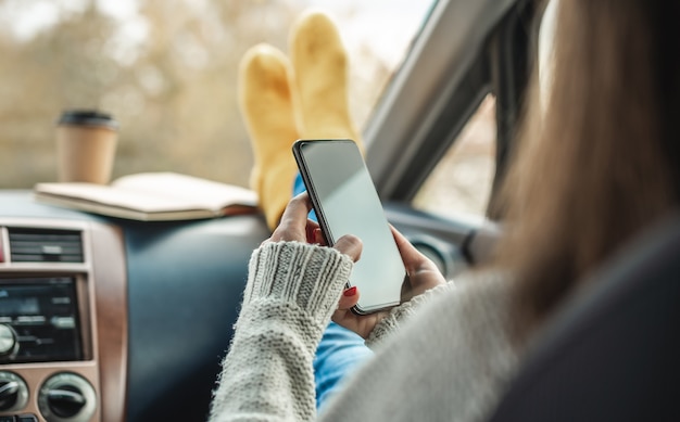 Photo woman in a car in warm socks is holding a mobile phone gadget and cup of coffee