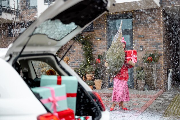 Woman carries christmas tree and gift box near a house