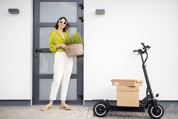 Woman carrying basket with herbs on a porch of her house with electric scooter and parcels nearby