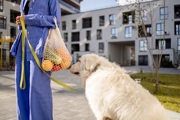 개와 함께 집으로 걸어가는 동안 신선한 식료품이 든 메쉬백을 들고 있는 여성