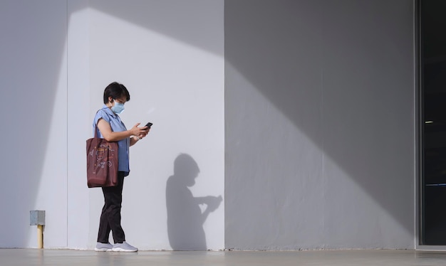 Woman carrying tote bag using smartphone while walking beside the entrance wall of supermarket