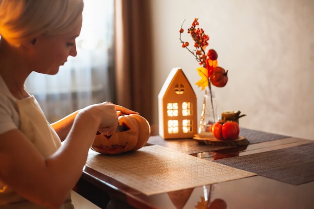 A woman carves a pumpkin for Halloween in a room with autumn decor and a lamp house. Cosy home and preparing for Halloween