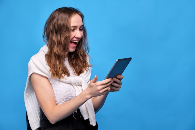 woman in casual clothes on blue sits and looks at the tablet in surprise and rejoicing.