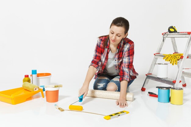 Woman in casual clothes sitting on floor with wallpaper rolls, paint roller, instruments for renovation apartment isolated on white background. Accessories for gluing, painting tools. Repair concept.