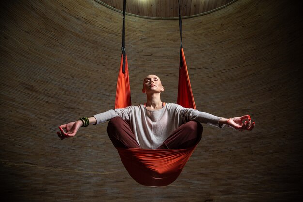 Woman in casual clothes sitting in orange hammock and putting her hands on the knees while meditating